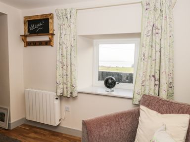 A window with floral curtains a small globe on the sill a wall-mounted radiator a brown sofa with cushions and a wooden framed welcome sign on the wall at The Granary Loft in Bardsea
