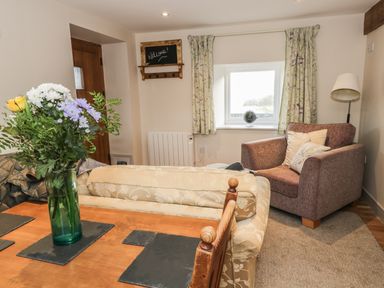 A sitting area with an armchair and cushions near a window with curtains a wooden table with a vase of flowers and placemats and a small chalkboard on the wall at The Granary Loft in Bardsea