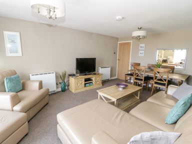 A living room with beige sofas a wooden coffee table a television on a wooden stand and a dining table with chairs at Harbour View in Cemaes Bay