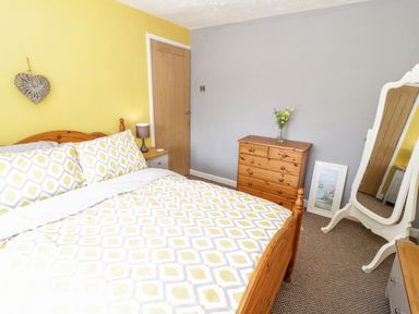 A bedroom with a wooden bed with patterned bedding a wooden dresser a standing mirror and a bedside table with a lamp at Harbour View in Cemaes Bay