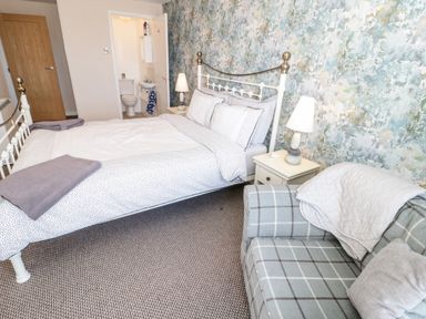 A bedroom with a bed table lamps a plaid armchair and an en suite bathroom at Harbour View in Cemaes Bay