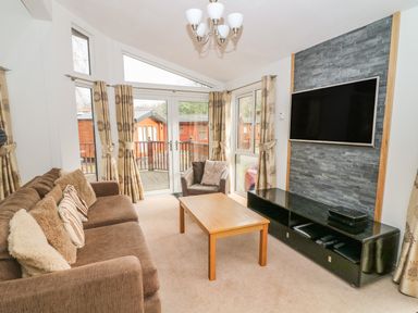 A living room with a brown sofa and armchair a wooden coffee table a wall-mounted TV and large windows with curtains at Coniston Number 9 in White Cross Bay