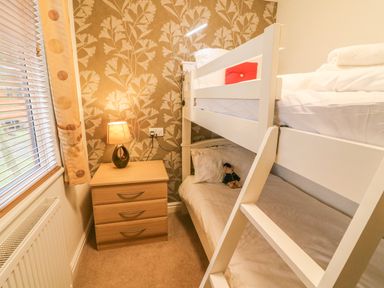 A small bedroom with a white bunk bed a wooden bedside table with a lamp and a window with blinds and curtains at Coniston Number 9 in White Cross Bay
