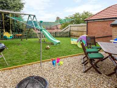A backyard with a swing set slide rotary clothesline patio table and chairs with cushions barbecue and children's toys at Tus Nua Cottage in Carhampton
