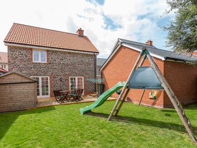 A backyard with a children's slide and swing set next to a stone house and a wooden shed at Tus Nua Cottage in Carhampton