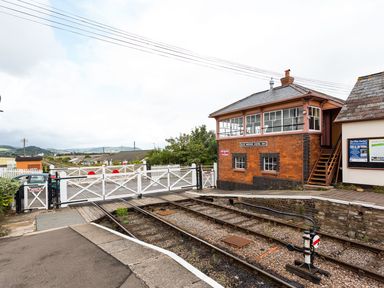 A railway crossing with closed white gates next to a brick signal box labeled Blue Anchor Signal Box at Tus Nua Cottage in Carhampton