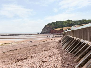A pebble beach with a concrete sea wall and people walking near cliffs at Tus Nua Cottage in Carhampton