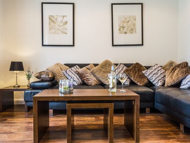 A living room with a black leather sofa with multiple cushions a wooden coffee table with a bottle and glass of white wine a small side table with a lamp and a vase with flowers and two framed artworks on the wall at Tus Nua Cottage in Carhampton