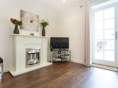 A room with a white fireplace decorated with two vases and a painting a TV on a glass stand and a door leading outside at Tus Nua Cottage in Carhampton