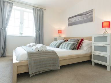 A bedroom with a wooden bed with pillows and folded towels a window with curtains and bedside tables with red lamps at Tus Nua Cottage in Carhampton
