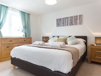 A bedroom with a bed with pillows and rolled towels a wooden dresser with a lamp and blue curtains at Tus Nua Cottage in Carhampton