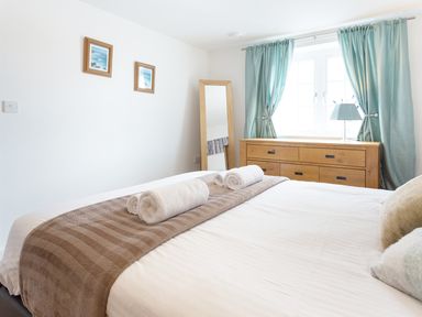 A bedroom with a bed covered with a brown throw and white towels on it next to a wooden dresser with a lamp and green curtains at Tus Nua Cottage in Carhampton