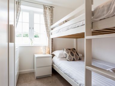 A bedroom with white bunk beds a bedside table with a lamp a window with curtains and a wardrobe at Tus Nua Cottage in Carhampton