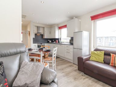 A kitchen with white cabinets and a refrigerator next to a dining table with chairs in a room with a brown leather sofa and a gray armchair at Jakanori in Staveley