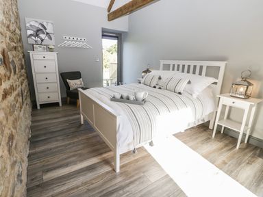 A bedroom with a white bed and striped bedding a gray chair a white chest of drawers a stone wall and wooden floor at Lowdale Barns West in Sleights