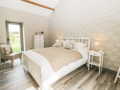 A bedroom with a white bed a beige blanket two nightstands with lamps a gray door a chair with a cushion and a white chest of drawers at Lowdale Barns East Sleights