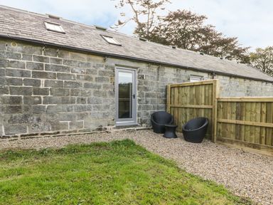 A stone building with a glass door and skylights with two black chairs and a small table on a gravel area behind a wooden fence at Lowdale Barns East in Sleights