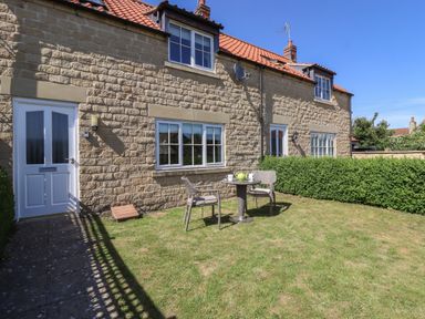 A stone house with a red tiled roof a white door and windows a small patio table with two chairs on a grassy lawn at 8 Pottergate Mews Helmsley