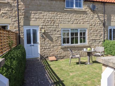 An outdoor garden area with two chairs and a table with a teapot and mugs in front of a stone house with a white door and windows at 8 Pottergate Mews in Helmsley