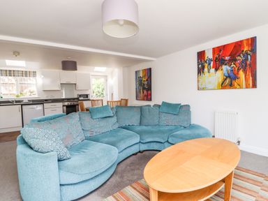 A living room with a curved blue sofa and wooden coffee table next to a kitchen with white cabinets and a dining table at 8 Pottergate Mews in Helmsley