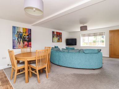 A living room with a round wooden dining table and four chairs a curved teal sofa a wall-mounted television and two colorful paintings at 8 Pottergate Mews in Helmsley