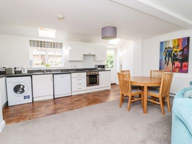 An open kitchen and dining area with white cabinets wooden dining table and chairs and a colorful painting on the wall at 8 Pottergate Mews in Helmsley