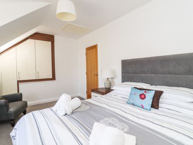 A bedroom with a striped bed, gray headboard, a nightstand with a lamp, a gray armchair, built-in wardrobes, and a wooden door at 8 Pottergate Mews Helmsley