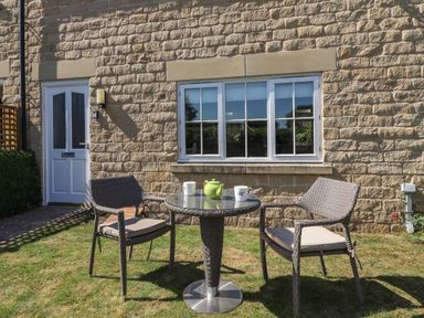 A small outdoor patio with two wicker chairs and a round table with a teapot and cups in front of a stone wall with a window and a white door at 8 Pottergate Mews in Helmsley