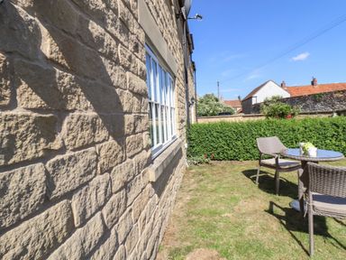 An outdoor patio area with a glass table and wicker chairs on grass next to a stone wall of a house with windows at 8 Pottergate Mews in Helmsley