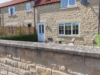 A stone house exterior with a white door and windows two chairs and a small table with a teapot outside behind a low stone wall at 8 Pottergate Mews in Helmsley