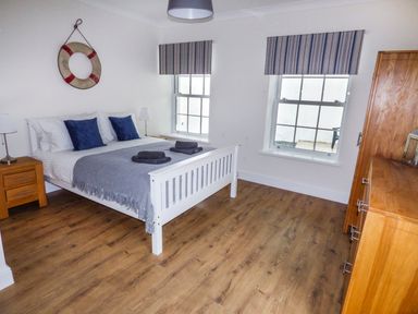 A bedroom with a bed with blue pillows a wooden chest of drawers and two windows at Harbour Beach at The Hideaway in Tenby