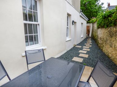 A narrow outdoor patio with a glass table and mesh chairs beside a white building with windows and a gravel path with stepping stones leading along a stone wall at Harbour Beach at The Hideaway in Tenby