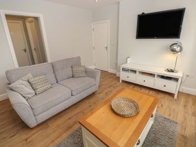 A living room with a gray sofa, wooden coffee table, white TV stand, wall-mounted TV, and a table lamp at Harbour Beach at The Hideaway in Tenby