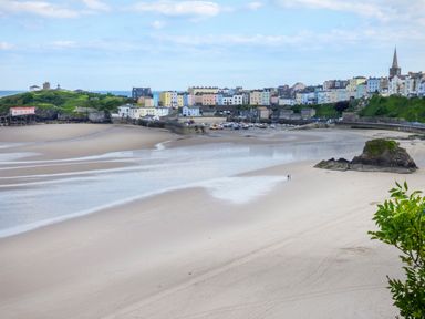 A sandy beach with a large rock and a small harbor with boats near colorful buildings and green hills at North Beach at The Hideaway in Tenby