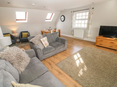 A living room with gray sofas a wooden dining table chairs a television on a wooden stand and windows at North Beach at The Hideaway in Tenby