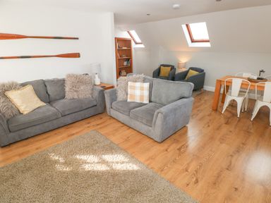 A living room with two gray sofas a beige rug wooden paddles on the wall two armchairs a wooden dining table with white chairs and wooden floors at North Beach at The Hideaway in Tenby