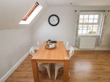 A dining area with a wooden table and four white chairs under a wall clock next to a window with patterned curtains at North Beach at The Hideaway in Tenby