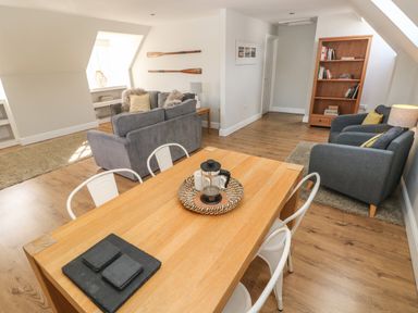 A living room with a wooden table with white chairs and gray sofas with cushions and a wooden bookshelf at North Beach at The Hideaway in Tenby