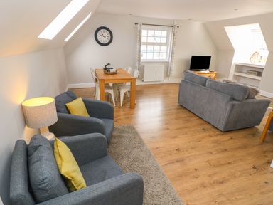 A living room with gray sofas yellow cushions a wooden dining table white chairs a TV on a wooden stand and a lamp at North Beach at The Hideaway in Tenby