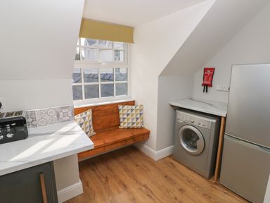 A kitchen corner with a wooden bench and cushions a washing machine a fridge and a toaster at North Beach at The Hideaway in Tenby