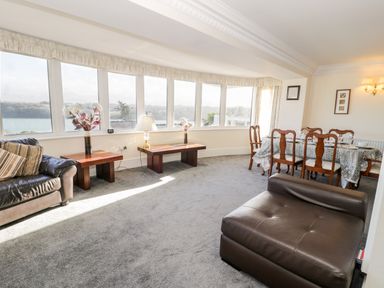 A living room with windows showing a water view two wooden tables a lamp and flowers a dining table set with chairs and a large brown leather chair at 11 Glyn Garth Mews Llandegfan near Beaumaris