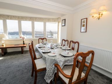 A dining room with a table set for six next to large windows and a bench with a lamp at 11 Glyn Garth Mews in Llandegfan near Beaumaris