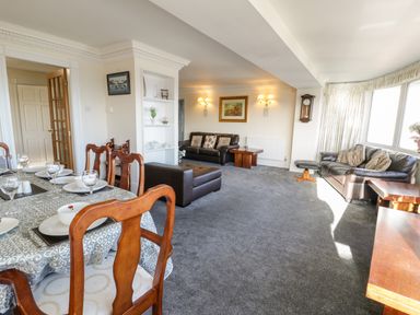 A living room and dining area with wooden chairs a dining table set with plates glasses leather sofas a wall clock and large windows at 11 Glyn Garth Mews in Llandegfan near Beaumaris