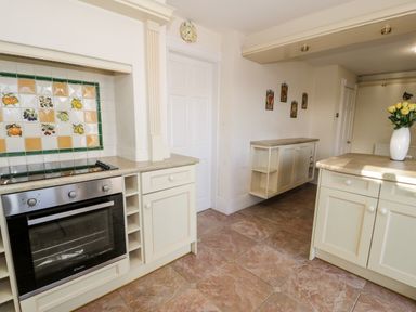 A kitchen with cream cabinets and tiled floor with a oven and decorated tiles on the wall at 11 Glyn Garth Mews in Llandegfan near Beaumaris