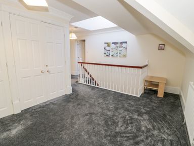 A hallway with grey carpet a staircase with white balustrade and wooden handrail a white double door closet and a wooden side table at 11 Glyn Garth Mews in Llandegfan near Beaumaris
