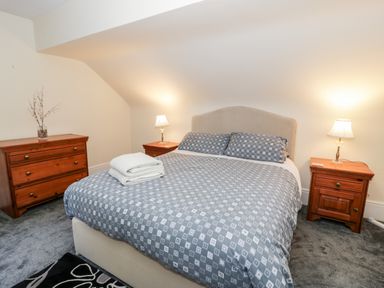 A bedroom with a double bed covered in a patterned grey duvet two bedside tables with lamps and a wooden chest of drawers at 11 Glyn Garth Mews in Llandegfan near Beaumaris