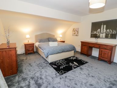 A bedroom with a double bed, bedside tables with lamps, a chest of drawers, a desk, and wall art at 11 Glyn Garth Mews in Llandegfan near Beaumaris