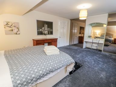 A bedroom with a bed, a wooden dresser, a wall mirror, a chair, and carpeted floor at 11 Glyn Garth Mews in Llandegfan near Beaumaris