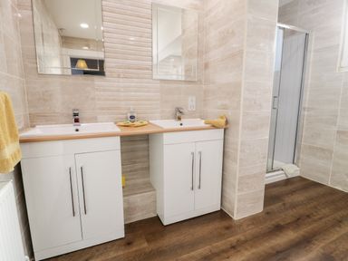 A bathroom with two sinks on white cabinets a shower area and beige tiled walls at 11 Glyn Garth Mews in Llandegfan near Beaumaris