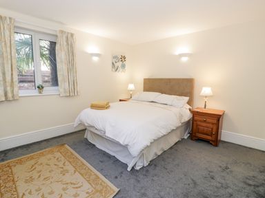 A bedroom with a bed white bedding two wooden bedside tables with lamps a window with curtains and a rug on the floor at 11 Glyn Garth Mews in Llandegfan near Beaumaris